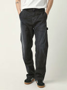 A person wearing black cargo-style pants with a white t-shirt and black sneakers, standing against a light gray studio background.