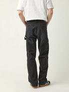 Rear view of a person wearing loose black cargo-style pants with a white T-shirt and black-and-white sneakers, standing against a plain light gray studio background.